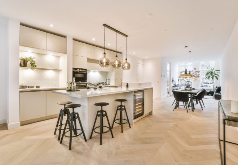 Open-concept kitchen with a marble island, four black bar stools, and hanging pendant lights, leading to a dining area.