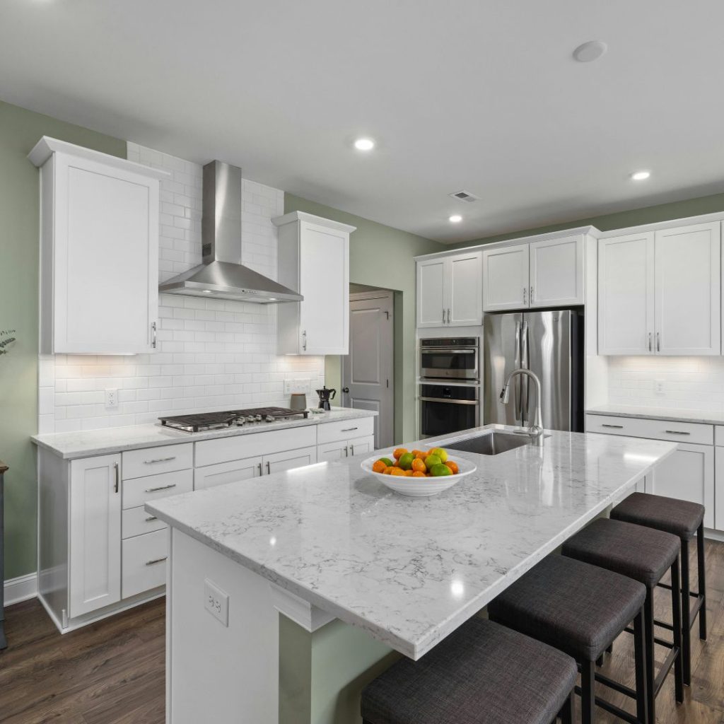 Modern white kitchen with marble island, fruit bowl, and stainless appliances. Green walls add color. Four dark stools line the island edge.