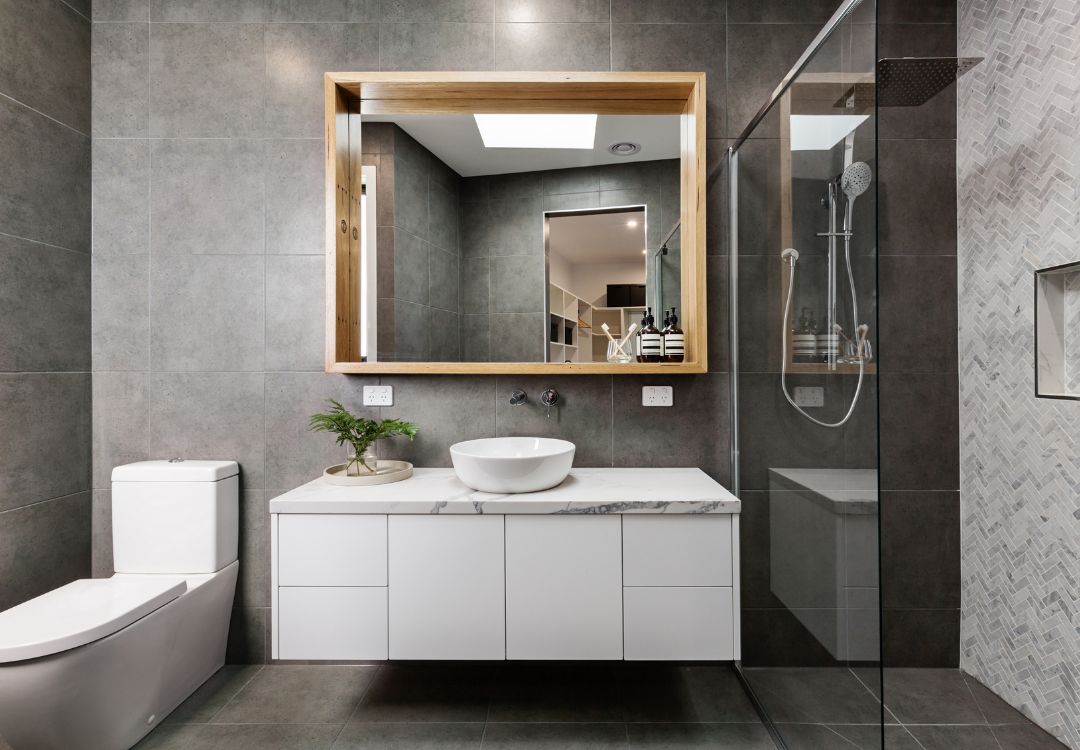 Modern bathroom with a white vanity, vessel sink, and a large wood-framed mirror against gray tiled walls, plus a glass-enclosed shower to the right.