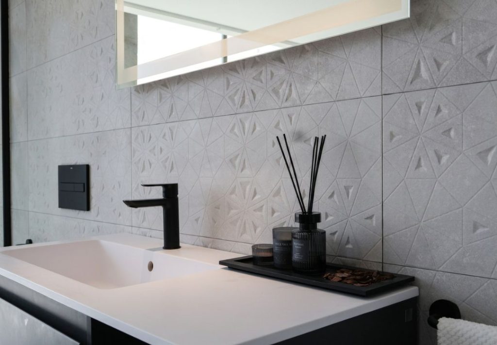 Modern bathroom sink with a black faucet, white countertop, and a black tray holding a diffuser and candles against a textured geometric tile wall.