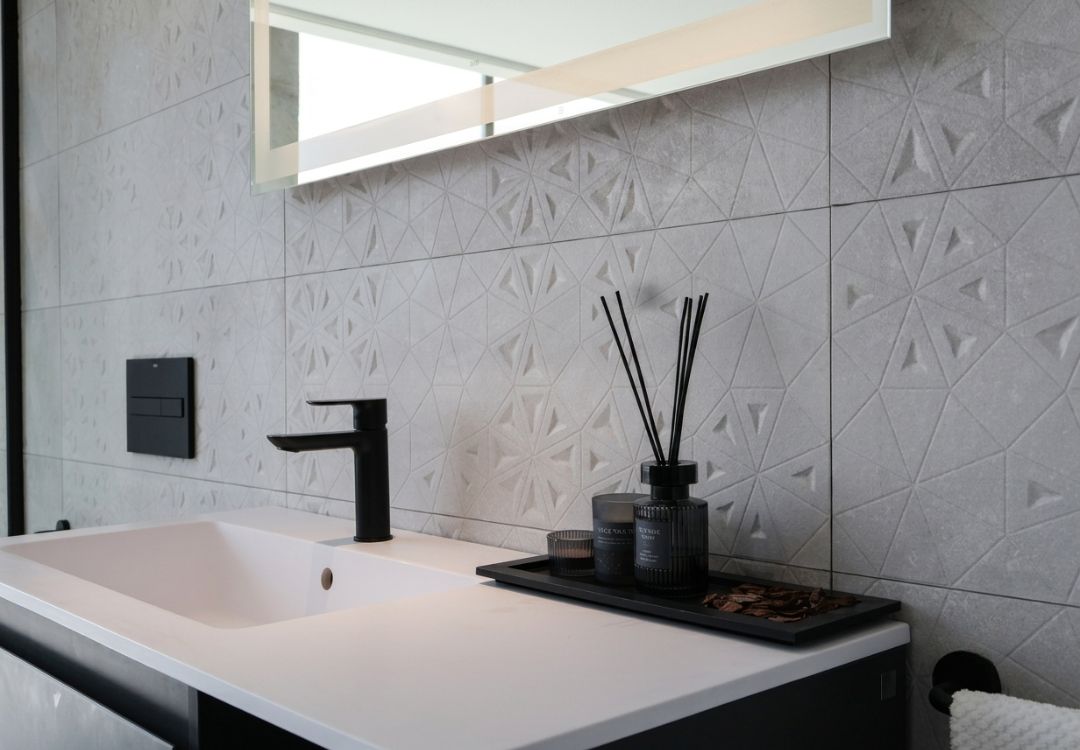 Modern bathroom sink with a black faucet, white countertop, and a black tray holding a diffuser and candles against a textured geometric tile wall.